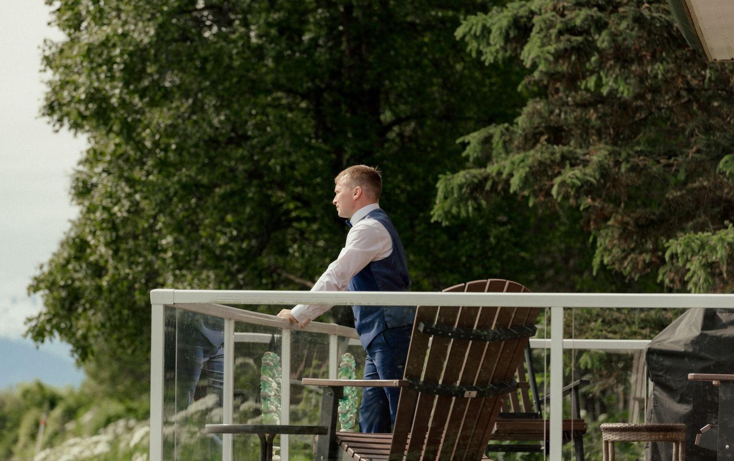 Man in vest stands on balcony overlooking lush green trees, gazing into the distance on a cloudy day.