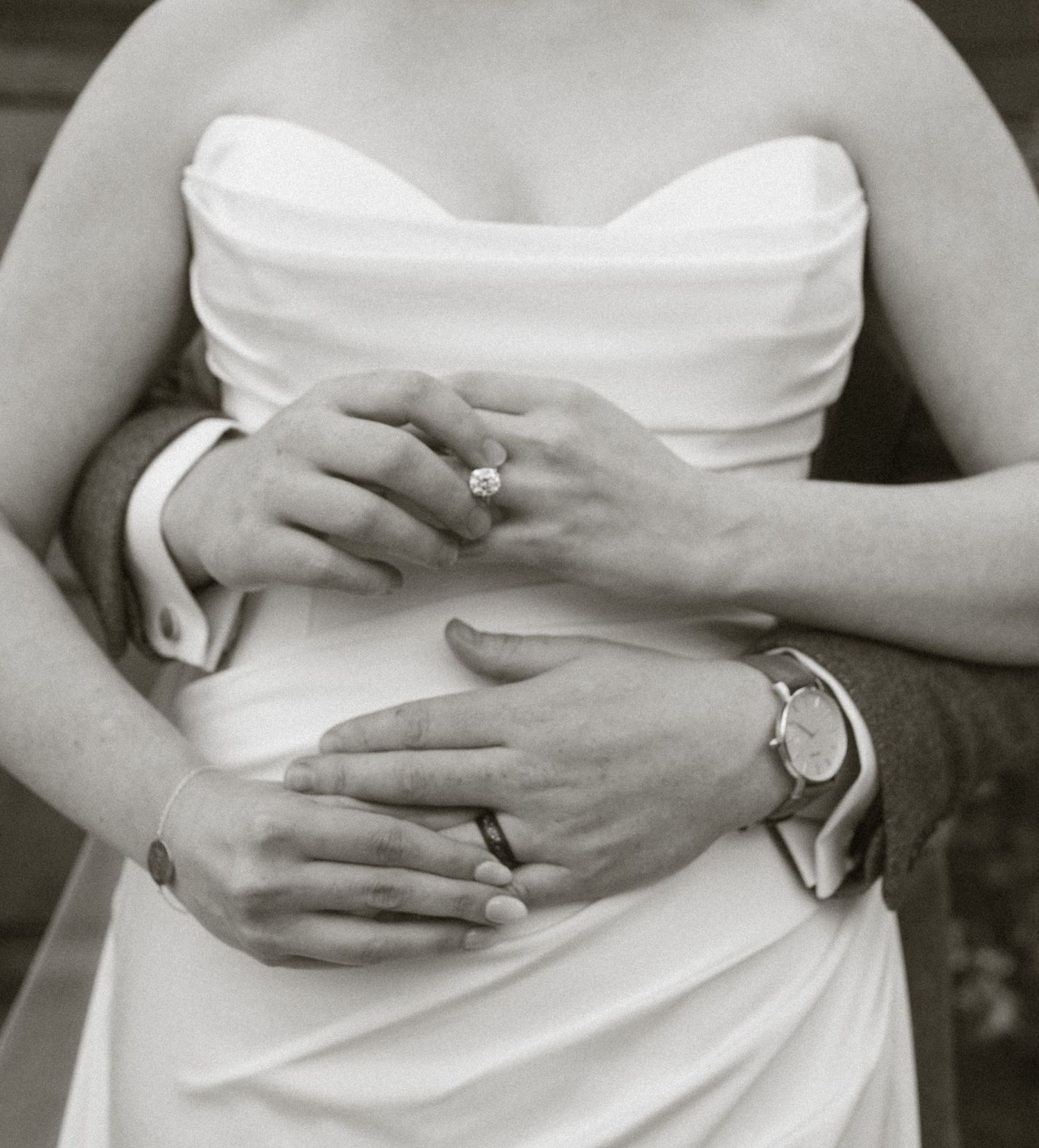 Black and white close-up of hands gently resting on a white dress fabric, showing a wedding ring and wristwatch.