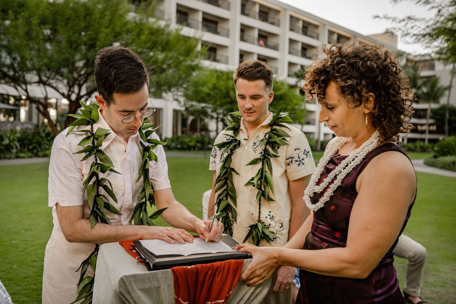 Two grooms in floral leis review wedding documents with officiant on Hawaiian resort lawn.