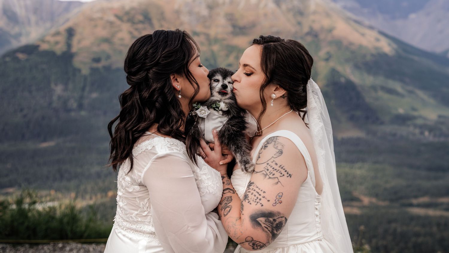 Two brides in white wedding dresses kiss their small dog outdoors with mountain scenery behind them.