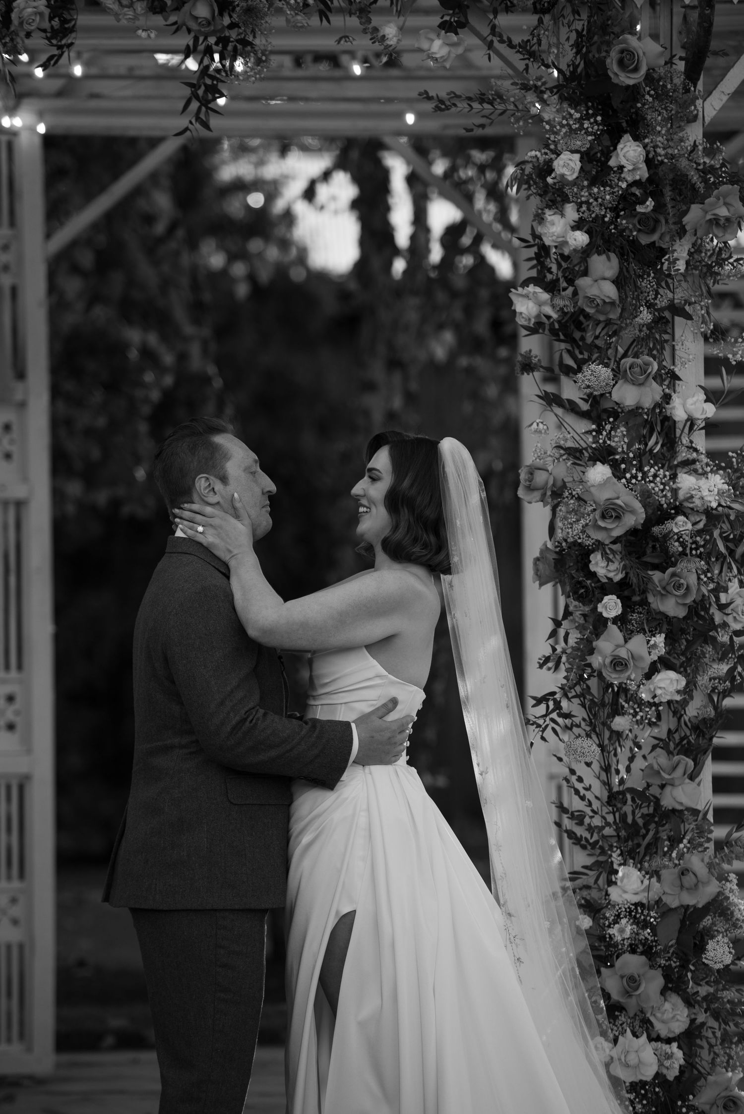 Black and white photo of bride and groom embracing under floral arch at outdoor wedding ceremony.