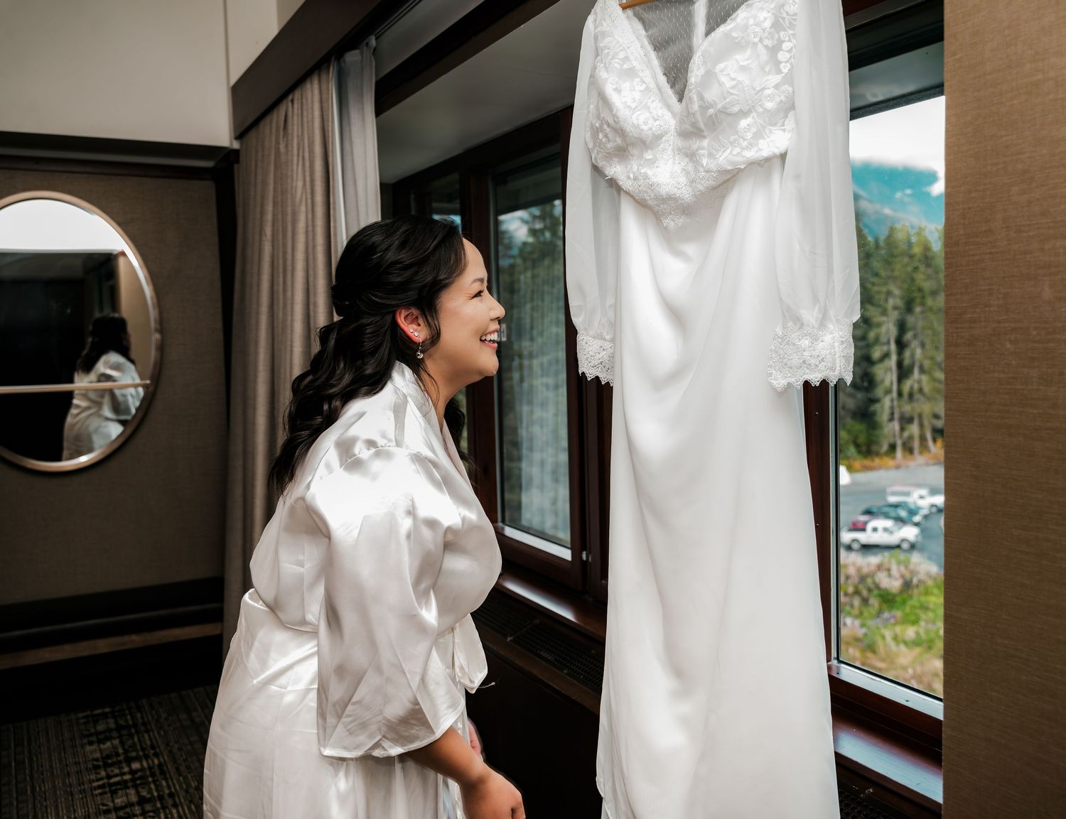 Bride in white satin robe smiling at her wedding dress hanging by a window in a hotel room.