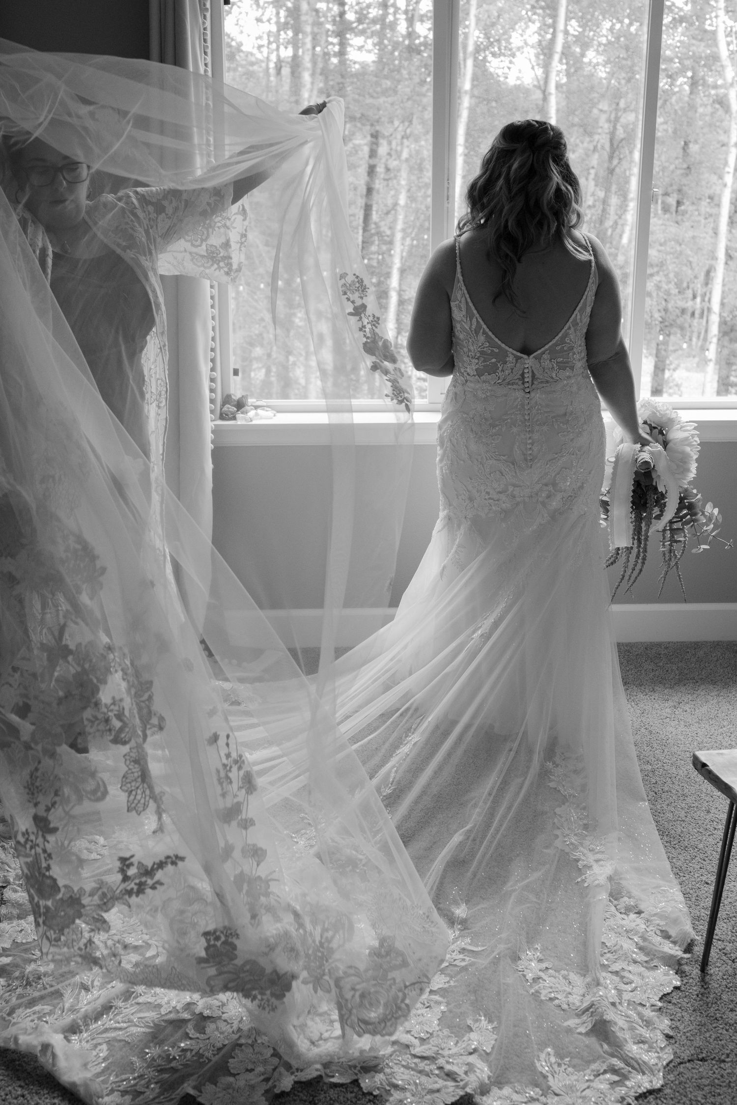 Black and white photo of a bride in a lace wedding gown with flowing veil, holding bouquet by a sunlit window.