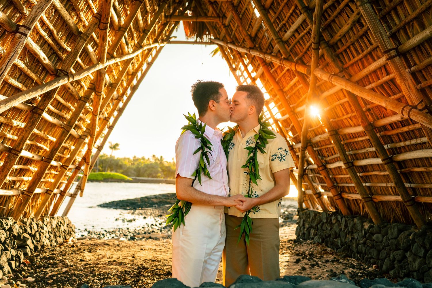 Two grooms sharing a kiss under a traditional Hawaiian thatched structure at golden sunset.