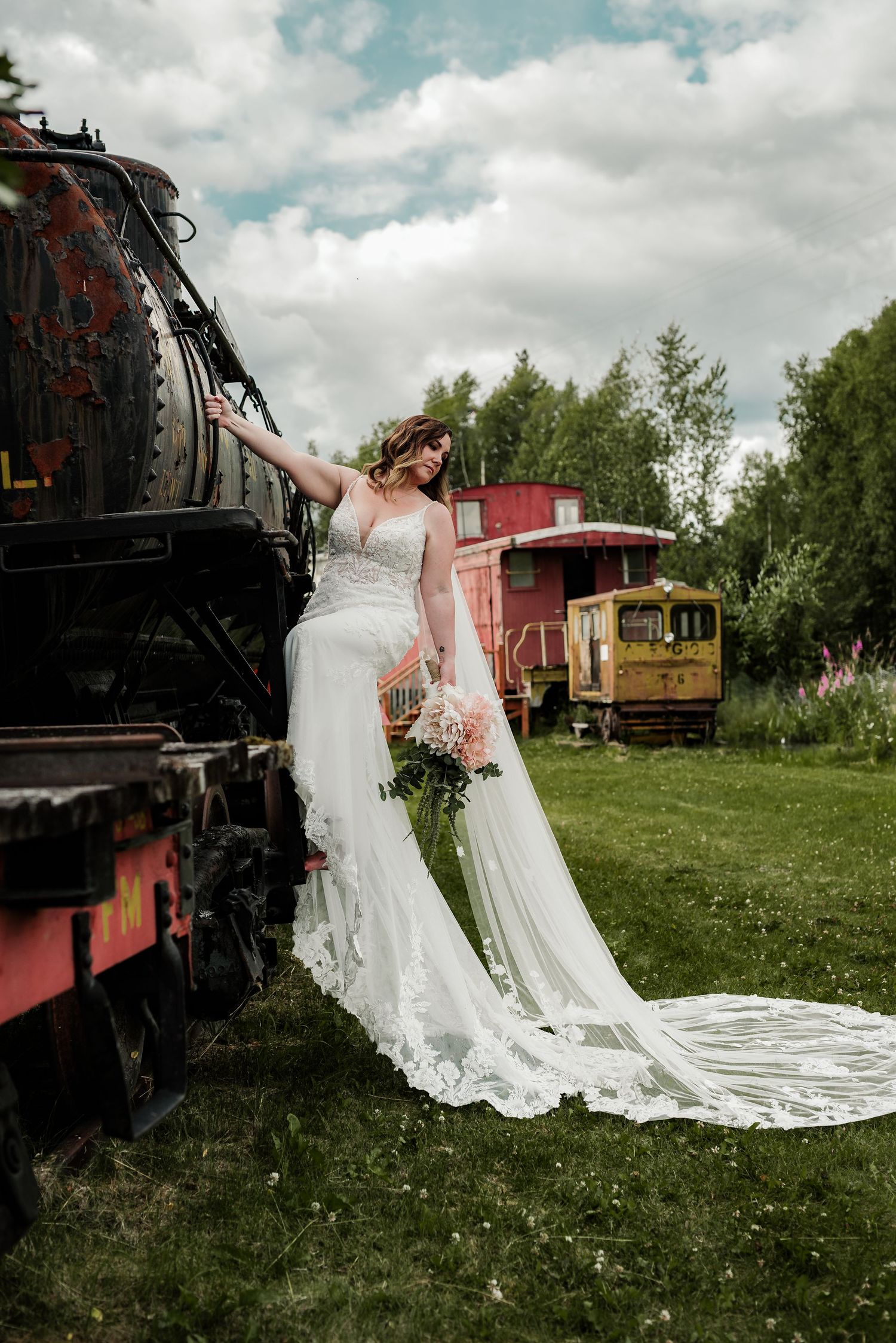 Bride in lace wedding gown poses beside vintage steam train holding pink bouquet with red caboose in background.