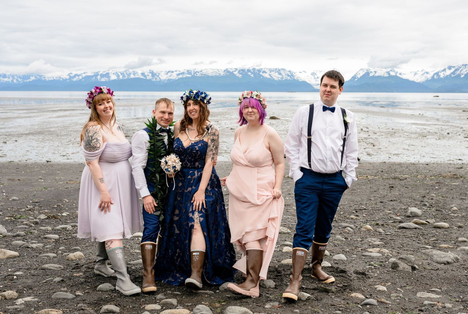wearing XtraTuffs Wedding party poses on rocky Alaskan beach with snow-capped mountains, bride in navy dress with floral crown.