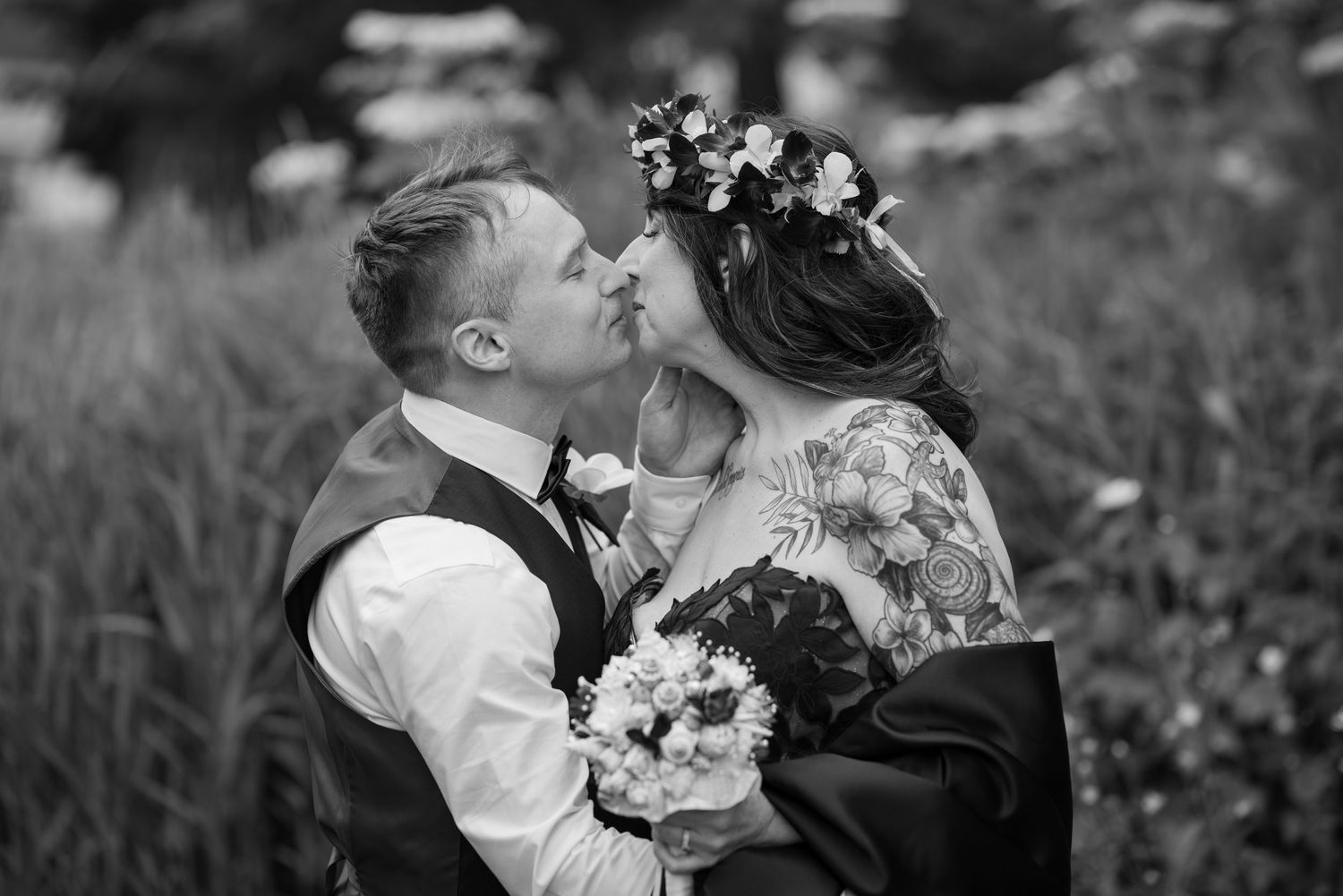Black and white wedding photo of a couple kissing, bride wearing floral crown and showing tattoos, groom in vest holding bouquet.