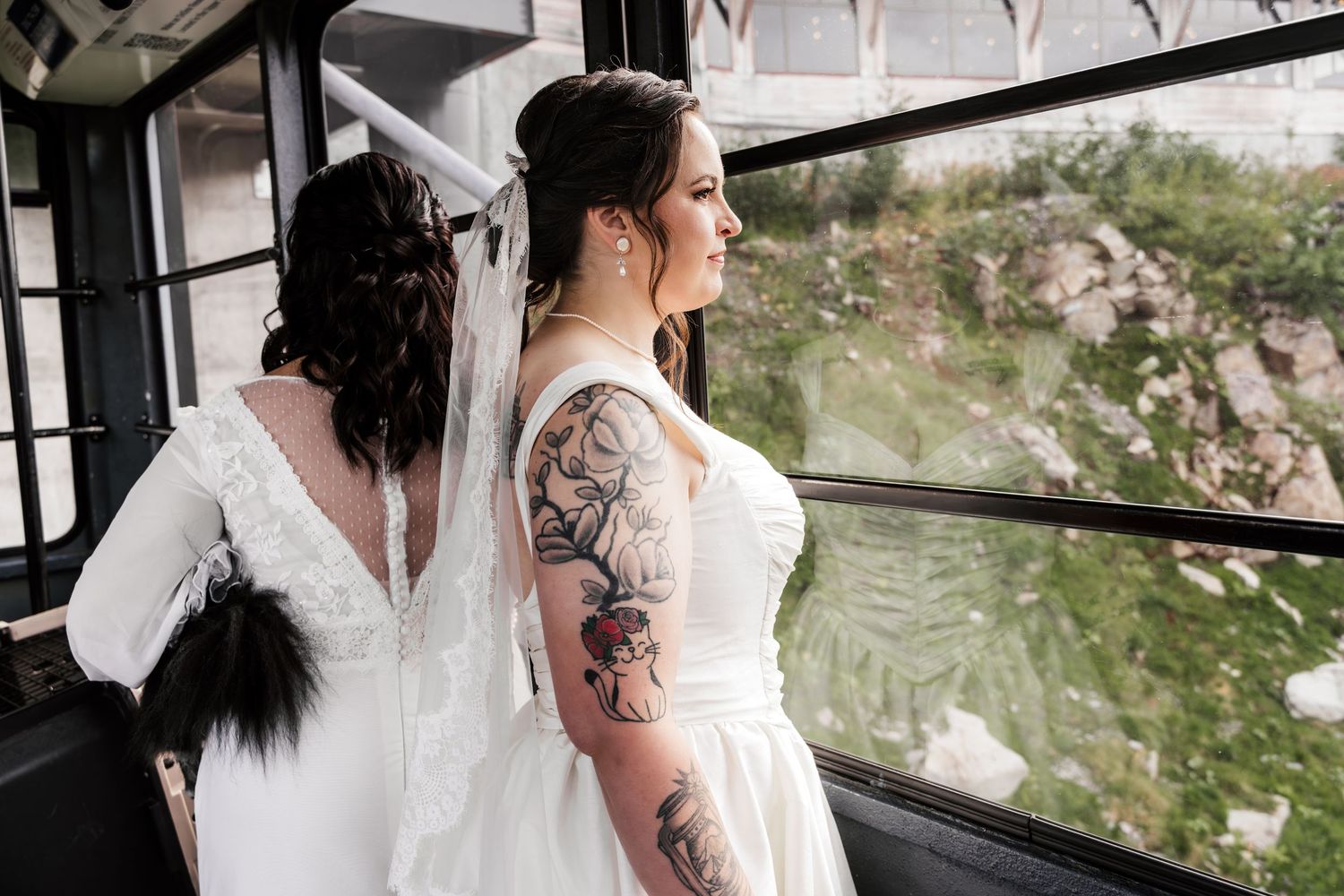 Two brides in white wedding gowns stand by a window, one with a veil and one with a floral tattoo sleeve.