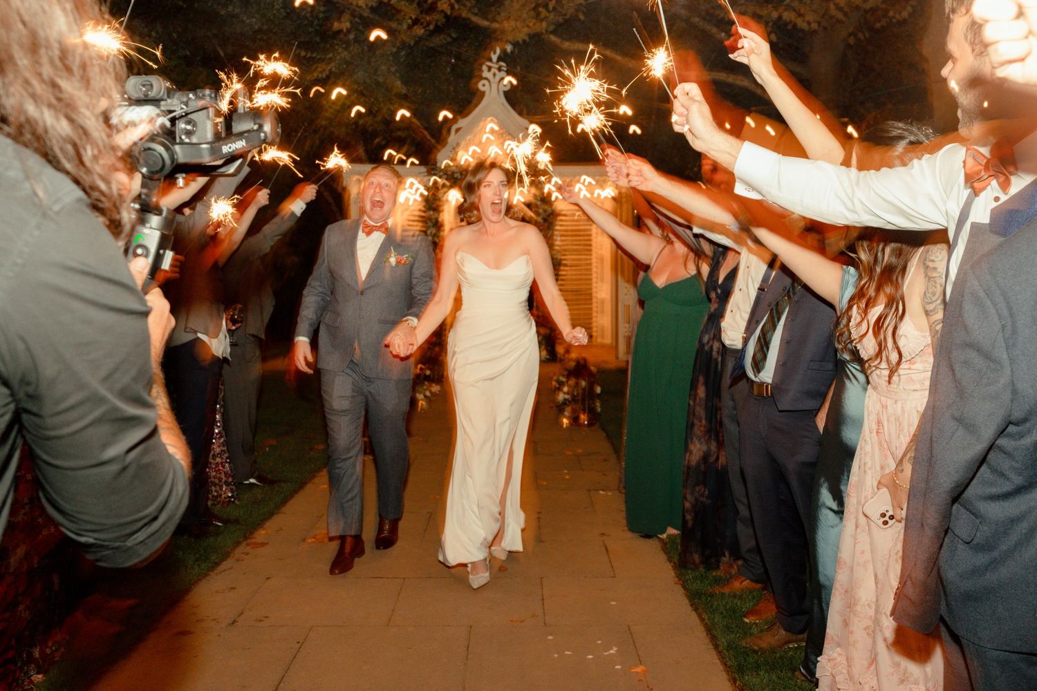 Bride and groom walk through sparkler tunnel with cheering guests at elegant outdoor wedding night send-off.