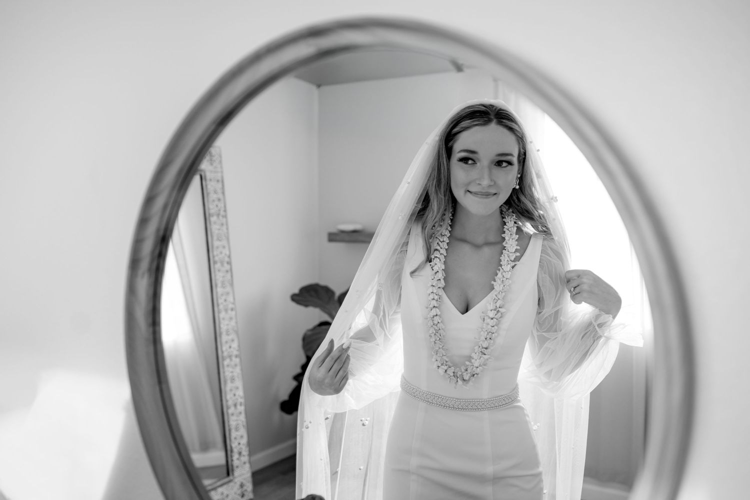 Black and white photo of a bride in a white dress and veil wearing a floral lei, reflected in a round mirror.