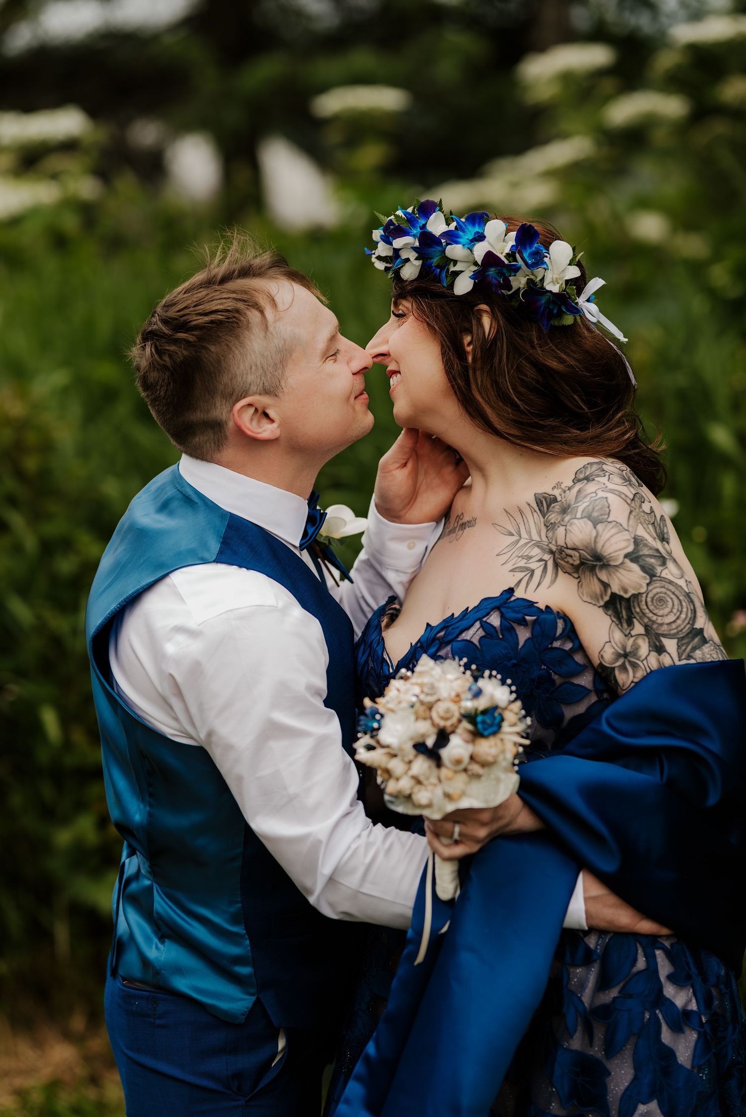 Bride in blue dress and floral crown shares romantic moment with groom in blue vest outdoors.