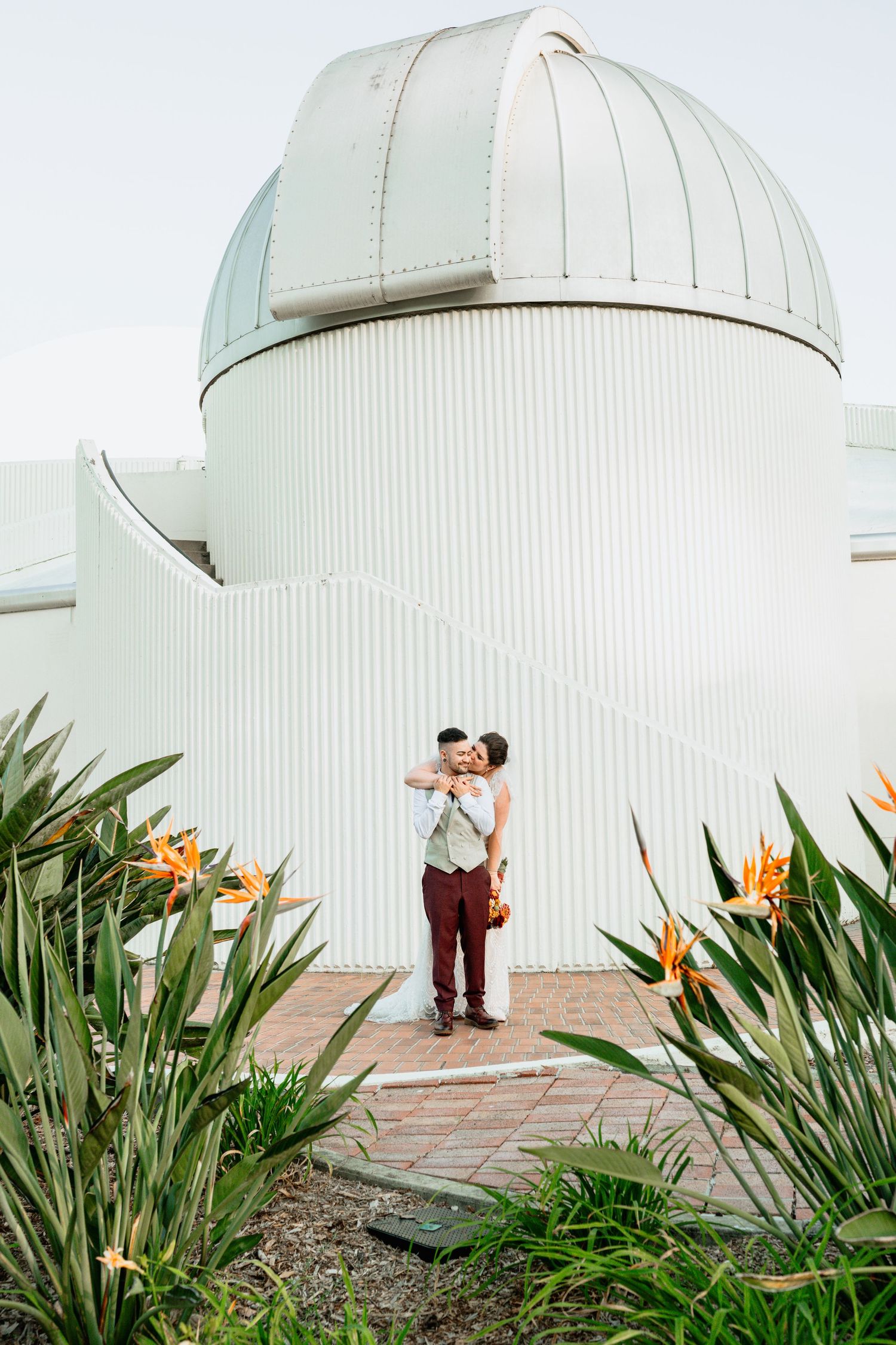 Couple embracing in front of a white observatory dome surrounded by bird of paradise flowers.
