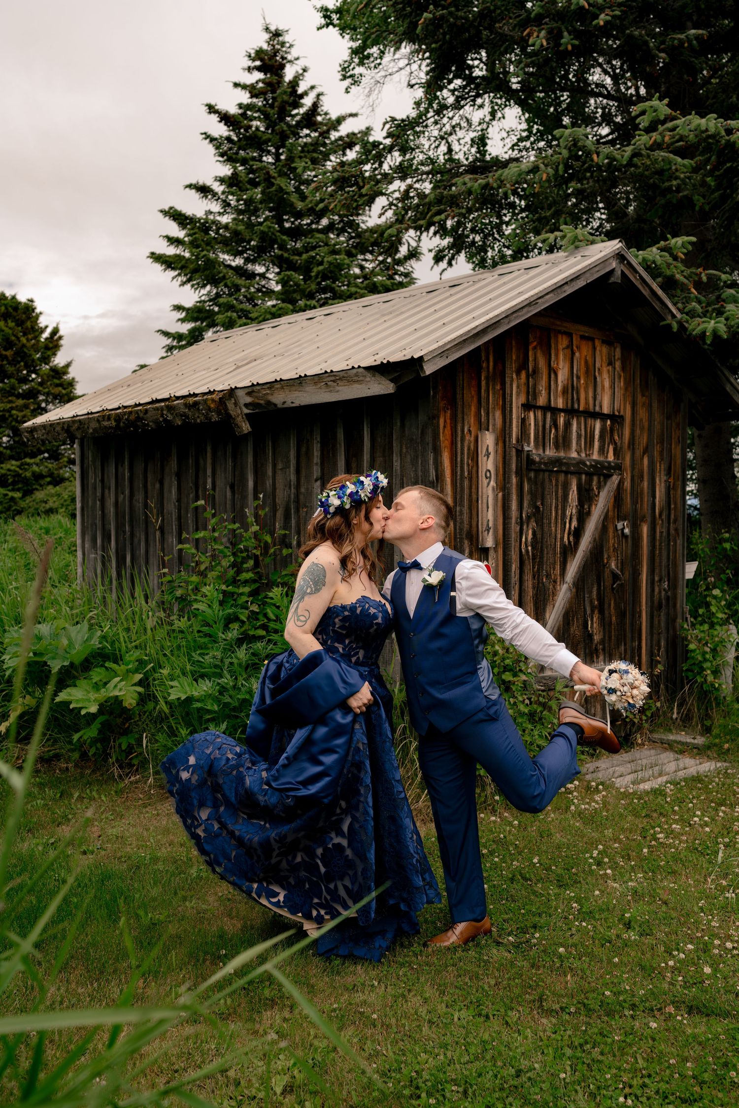 Couple kissing in matching blue wedding attire before rustic wooden shed, bride in floral crown holding bouquet outdoors.