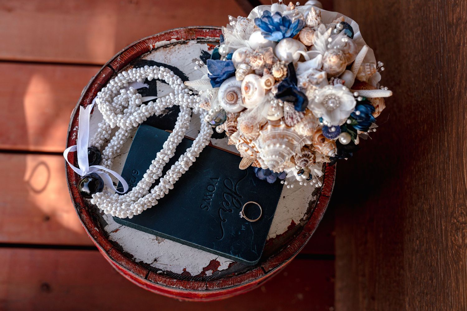 Round box with pearl necklace, wedding rings, blue flowers, and seashell bouquet on wooden surface in Homer Alaska