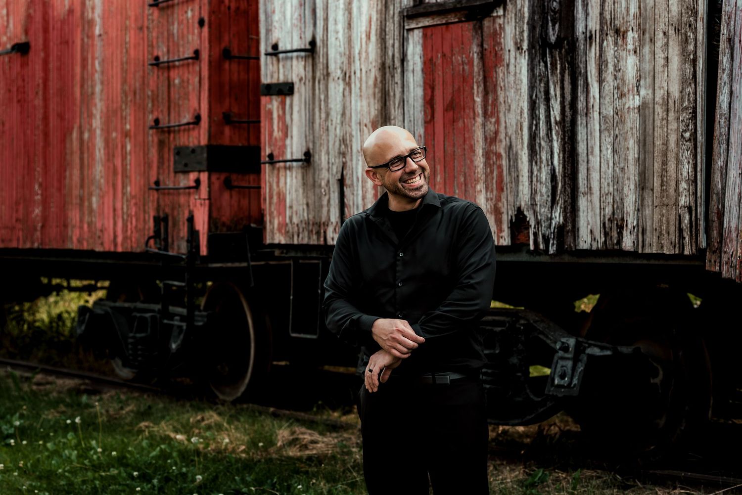 Bald man with glasses in black outfit smiling beside a vintage red wooden train car outdoors.