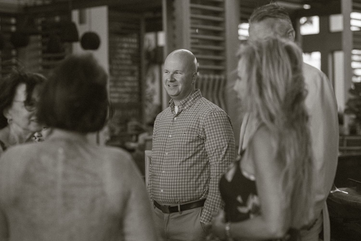 Black and white photo of smiling man in plaid shirt socializing with guests at an indoor gathering.