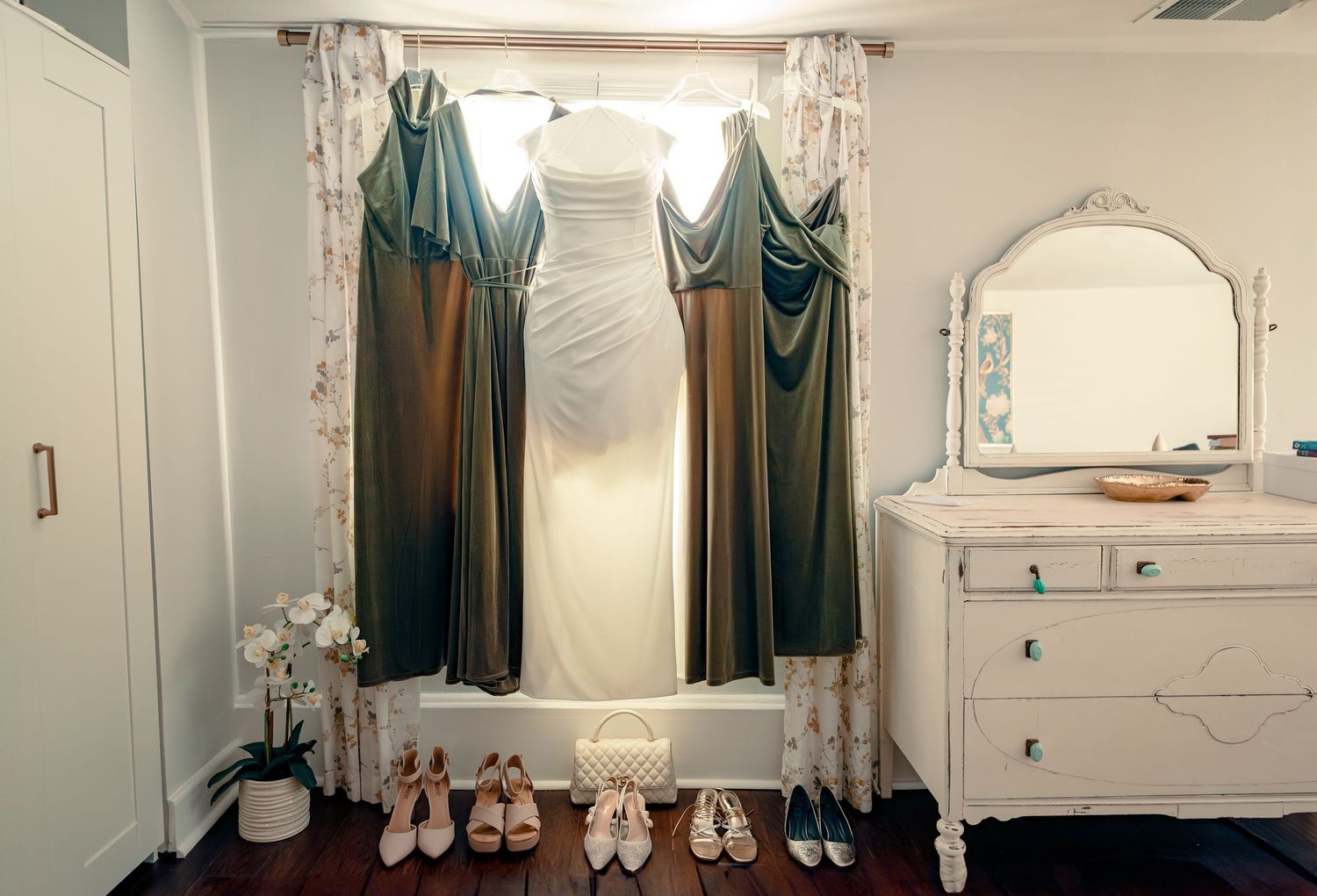 Elegant wedding dress displayed in window with green drapes, shoes arranged on dark hardwood floor, vintage white dresser.