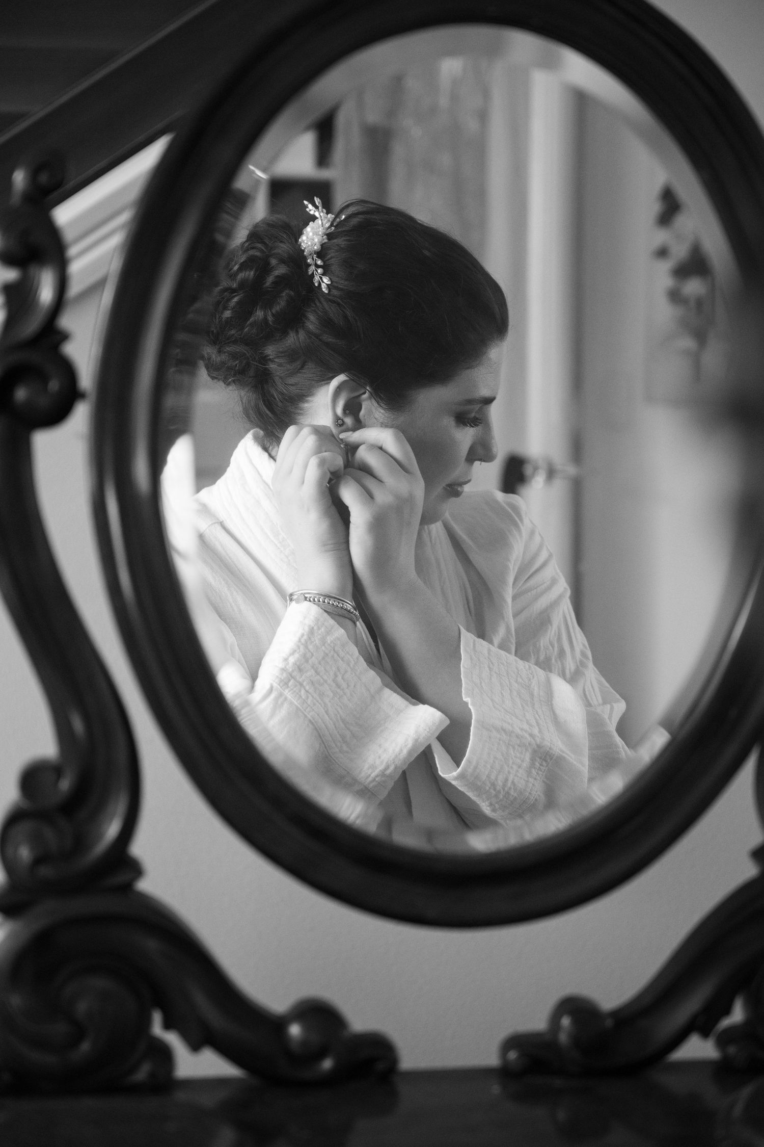 Black and white photo of a bride in a robe putting on earrings, reflected in an ornate oval mirror.
