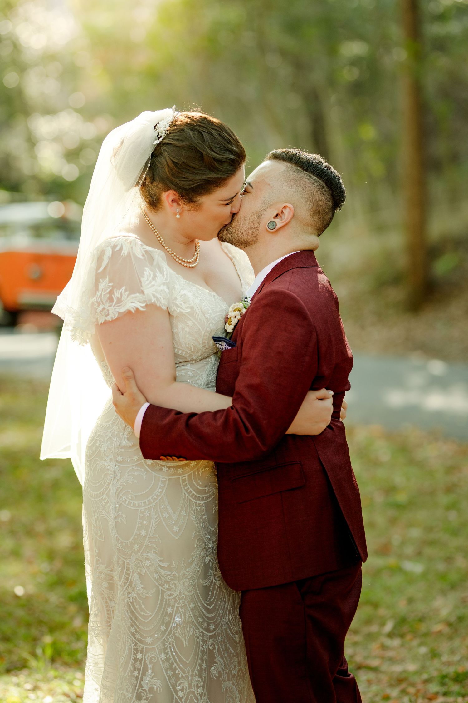 Bride in lace gown and groom in burgundy suit share a romantic kiss outdoors at their wedding ceremony.