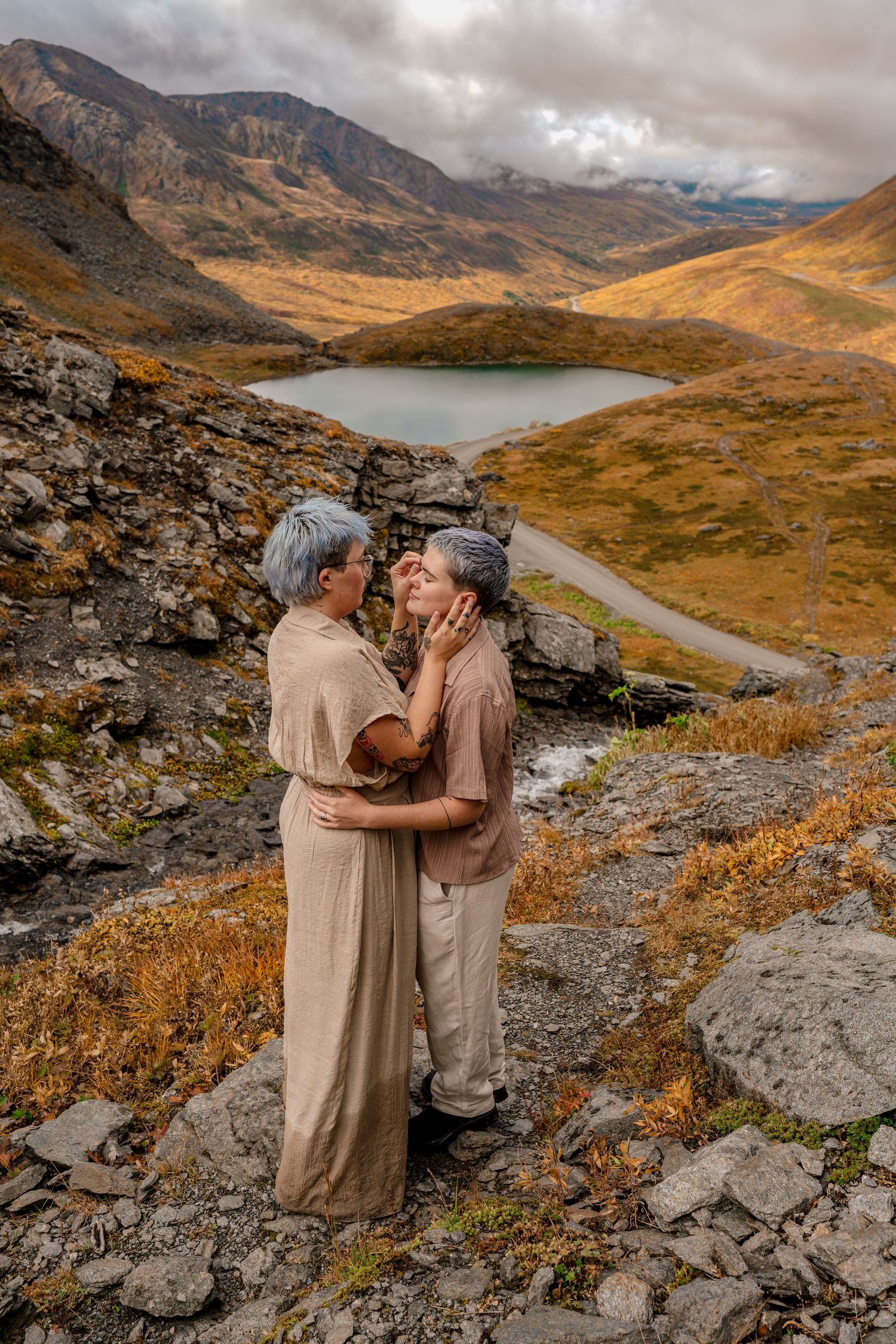 Two women with gray hair embrace romantically on a rocky mountain overlook with an alpine lake and golden autumn valley below.