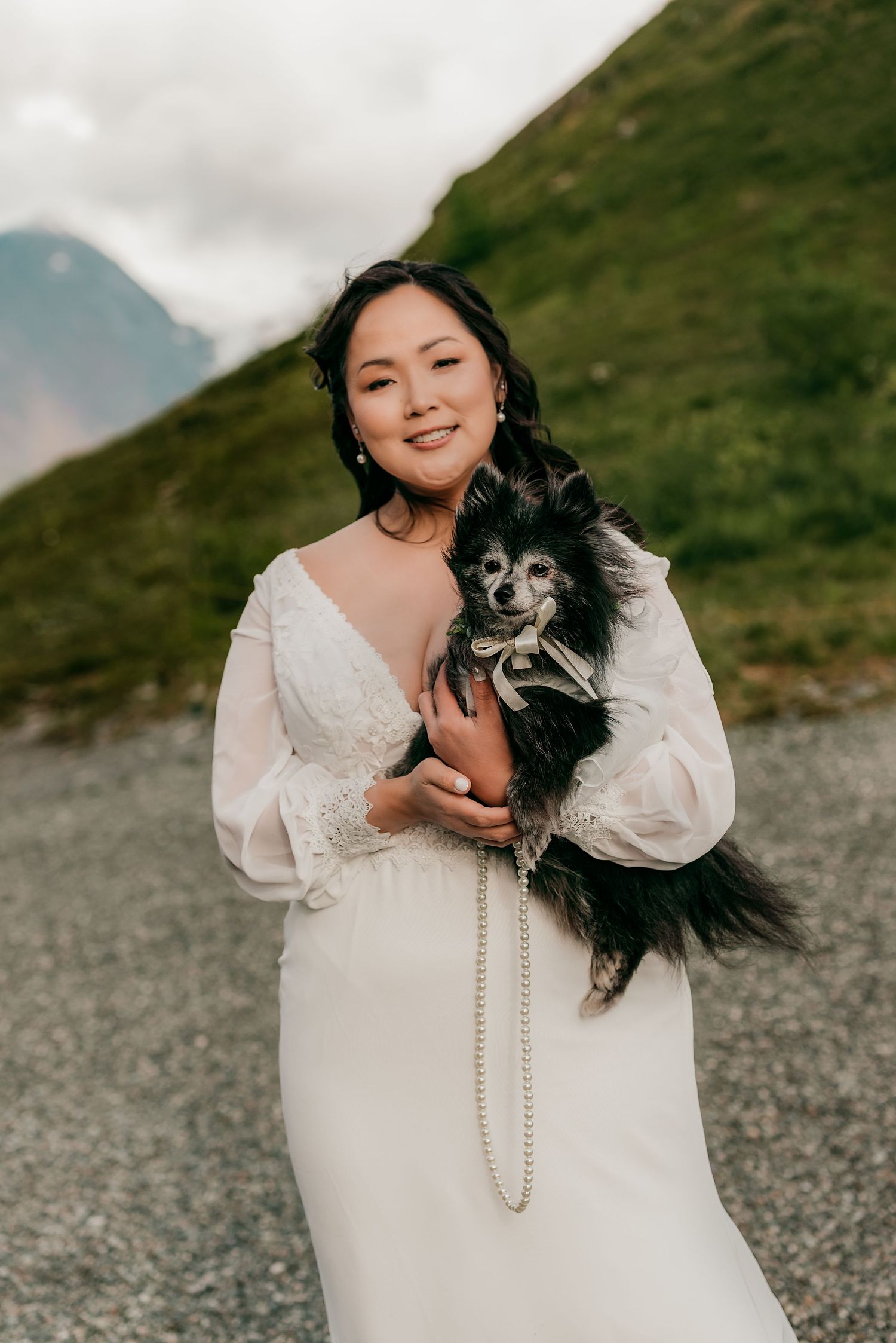 Bride in white lace gown holds a black and white Pomeranian with pearl bow, posing in a mountain landscape.