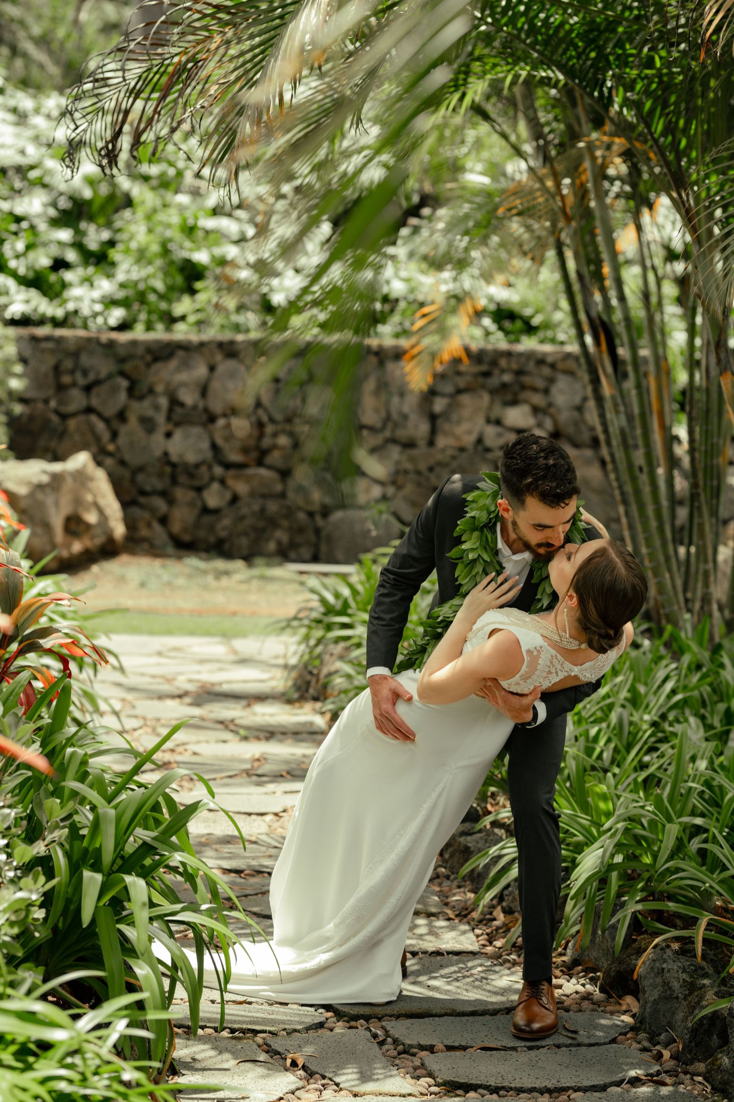 Intimate wedding moment captured in a tropical garden setting with stone wall and palm fronds.