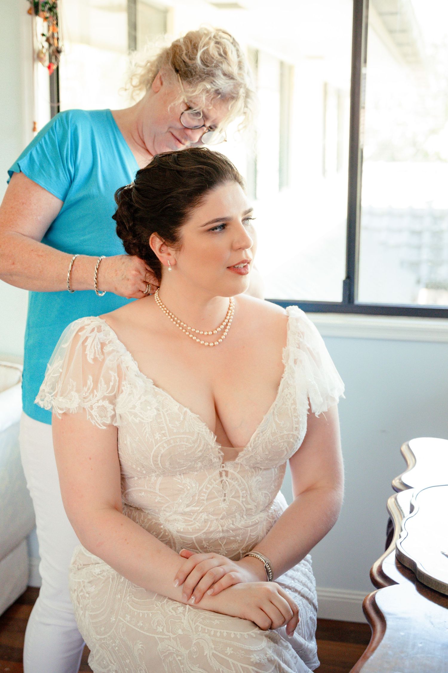 Bride in lace wedding gown with pearl necklace gets hair styled by older woman in blue top before wedding ceremony.