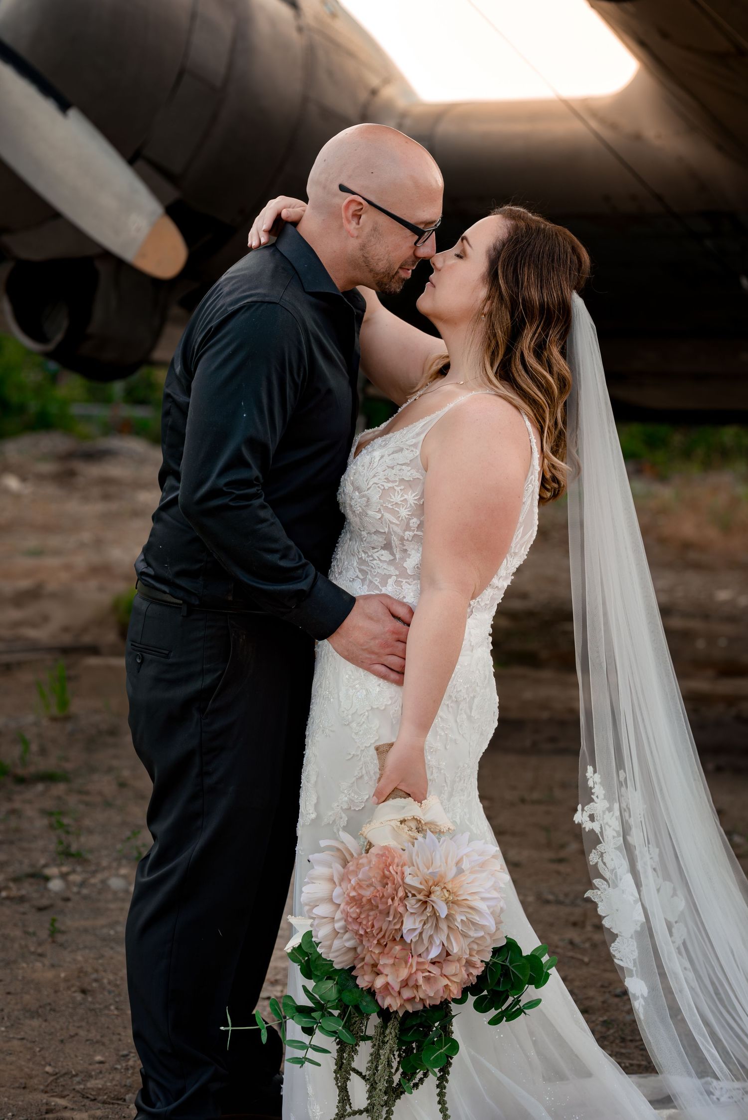 Bride in lace gown with veil and groom in black suit share romantic moment near vintage aircraft in Wasilla Alaska