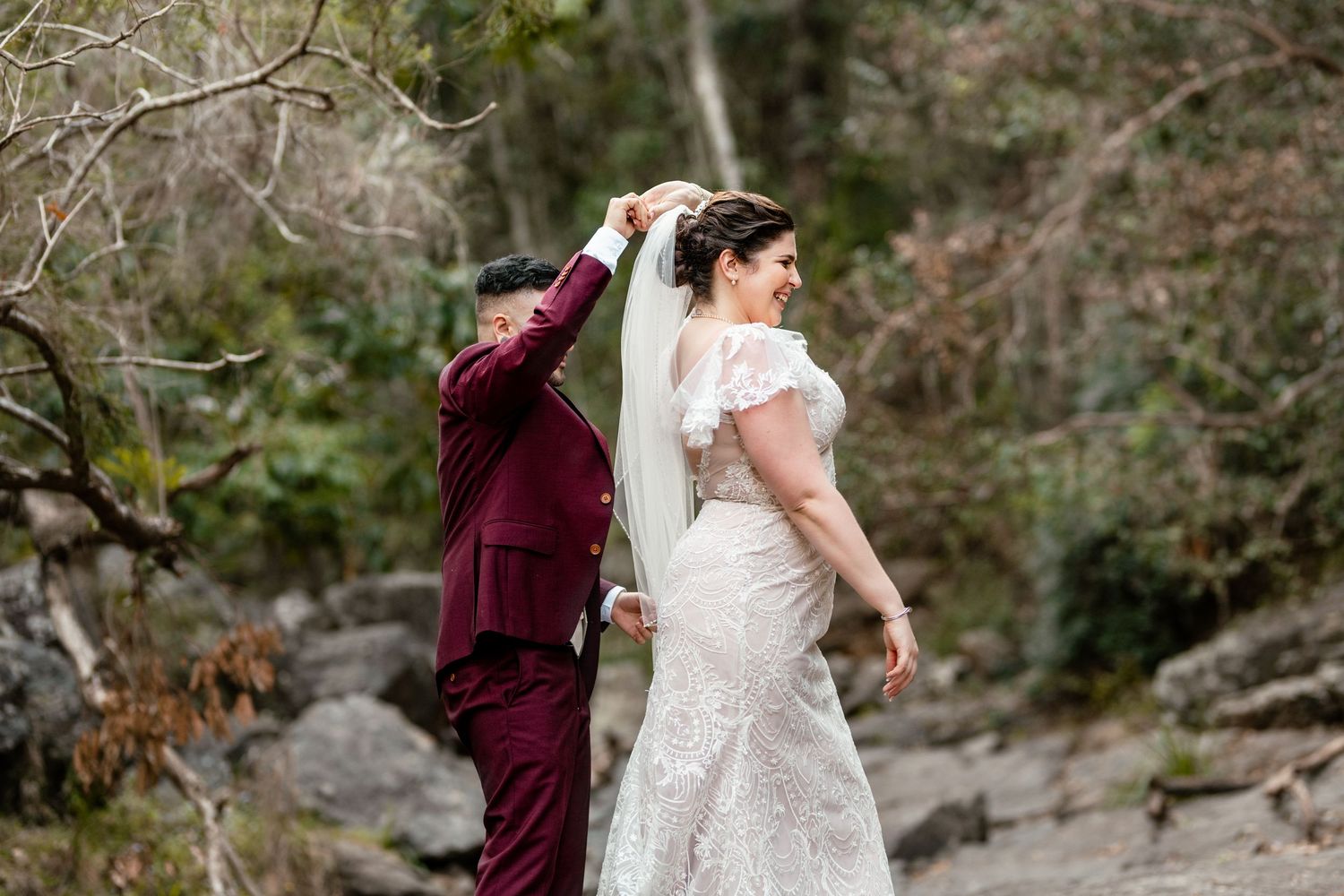 A bride in a lace gown and veil stands with a partner in a burgundy suit on a rocky outdoor wedding setting.