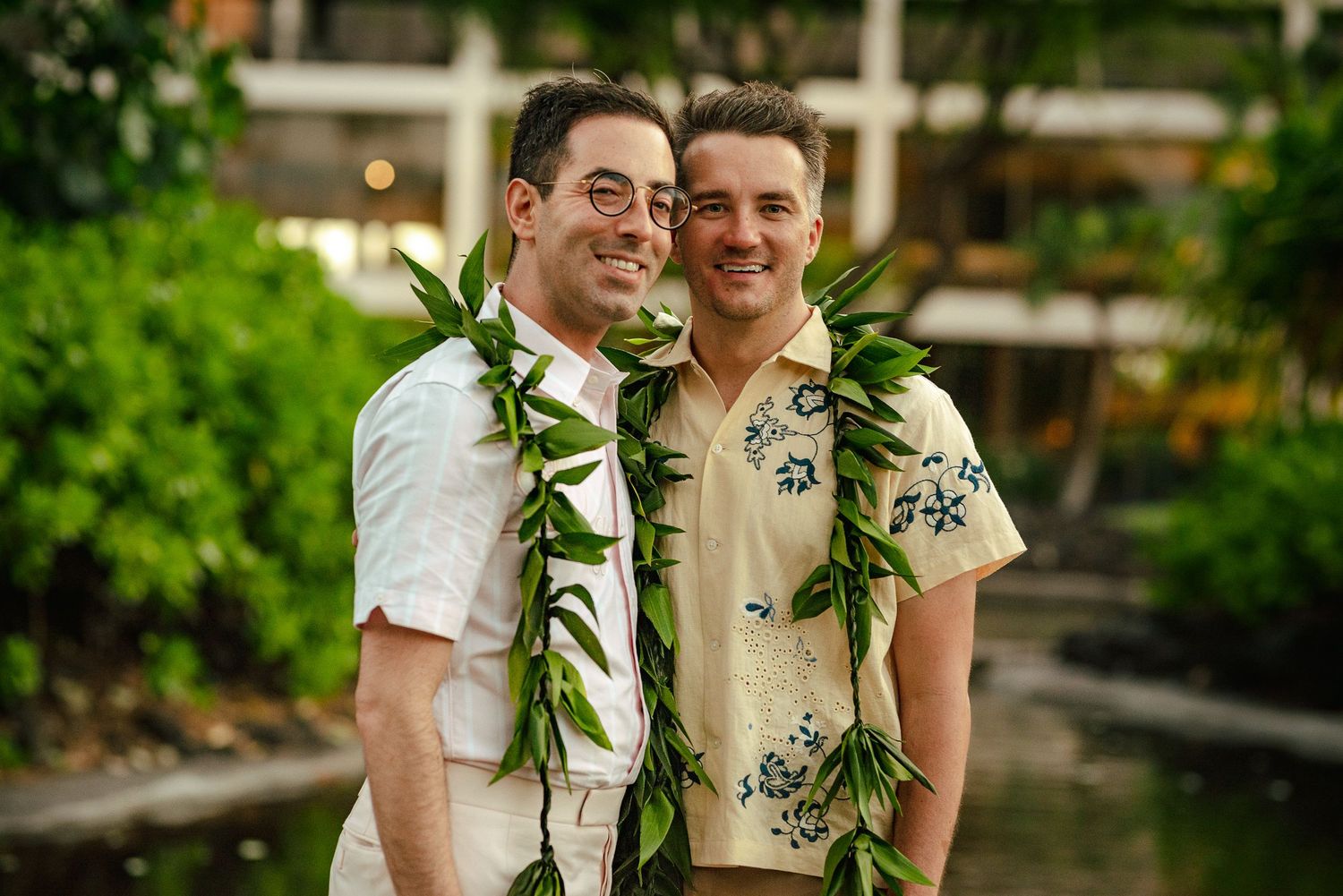 Two smiling men wearing Hawaiian leis pose together in a lush tropical garden setting.