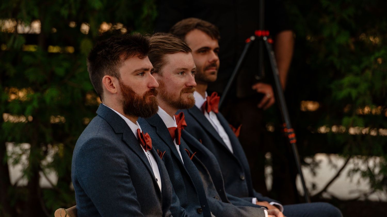 Three groomsmen in navy suits with red boutonnieres seated outdoors at a wedding ceremony.