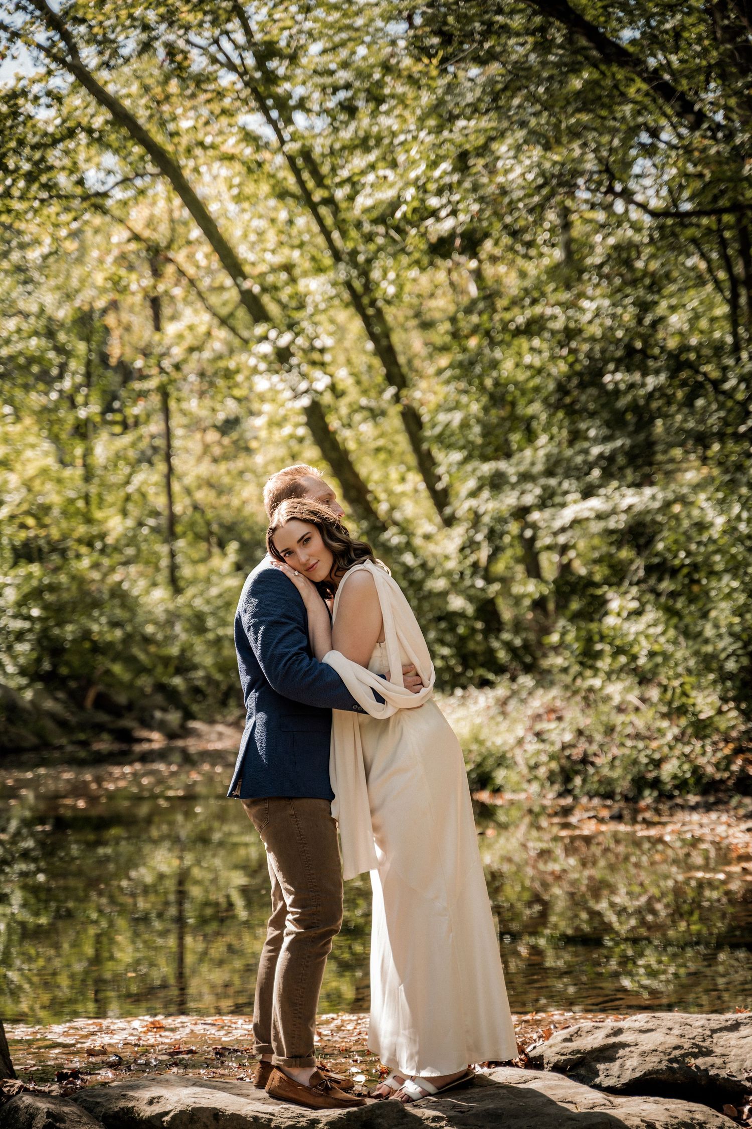 A couple embraces in a lush forest setting, the bride in a white gown and groom in a navy blazer, near a serene pond as they embrace.