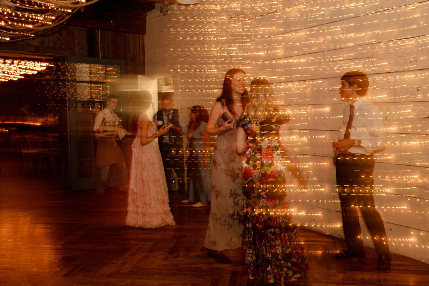 Elegant wedding reception with guests dancing near a glowing fairy light wall backdrop on a warm wooden floor.