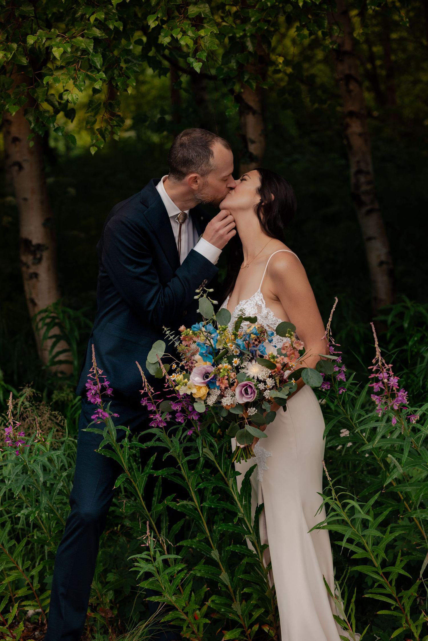Bride in sleek white dress holding colorful bouquet shares intimate moment with groom among purple wildflowers.