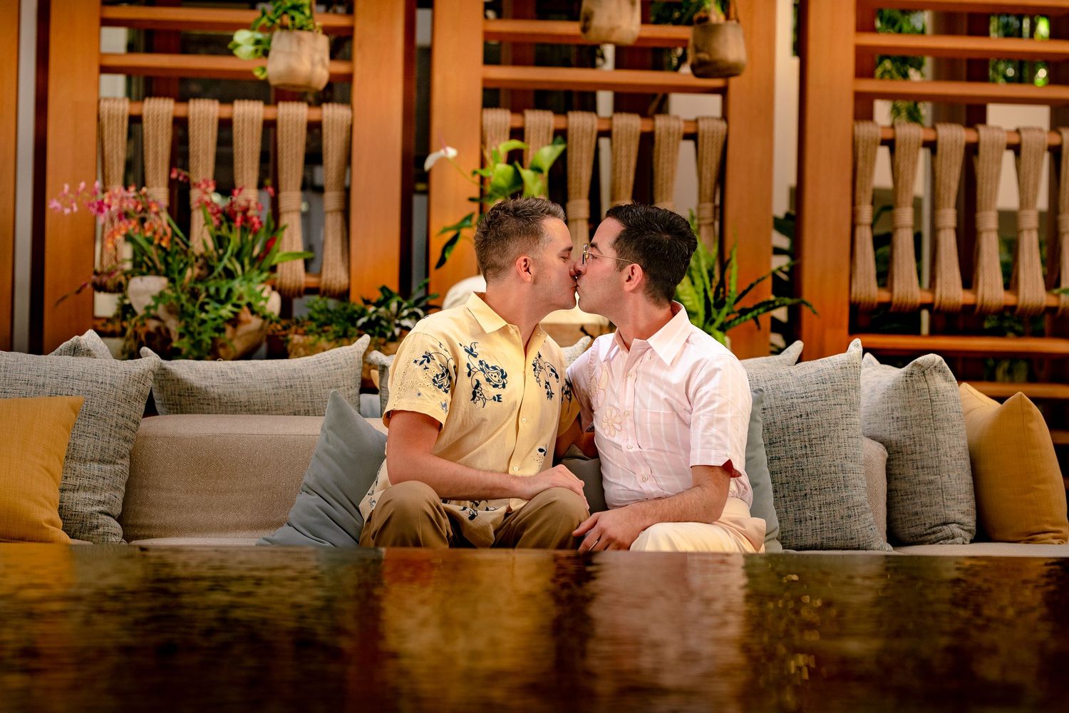 Two men sharing a romantic kiss on a couch in a tropical resort lounge with lush greenery decor.