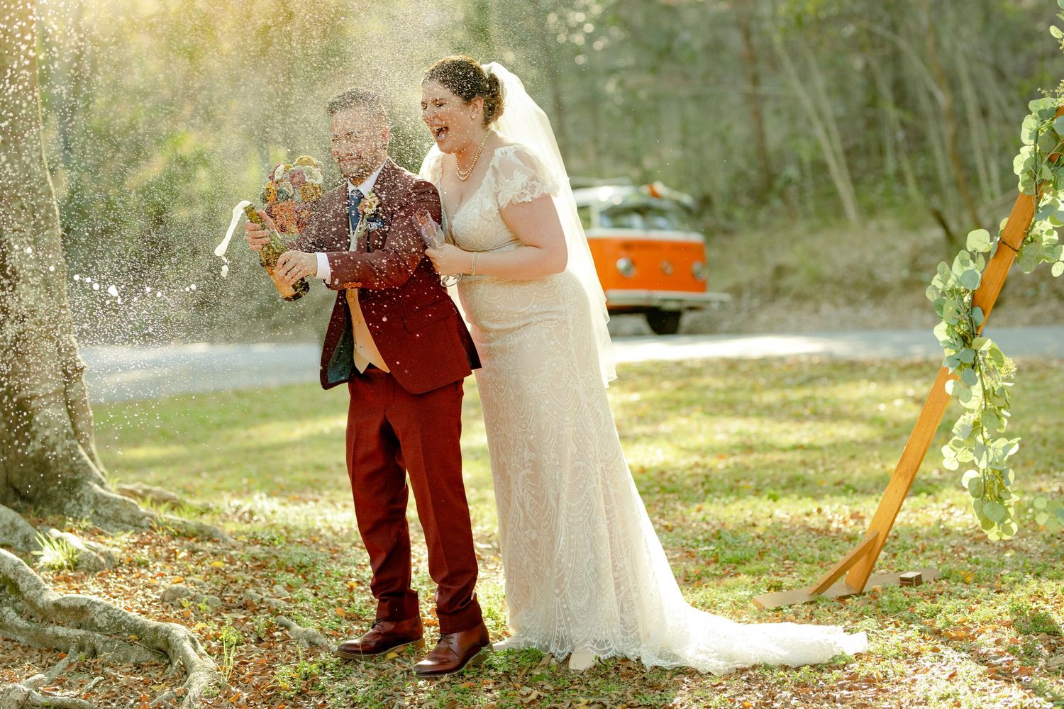 Joyful bride and groom in red suit celebrating with champagne spray outdoors near a vintage orange VW bus on their wedding day.