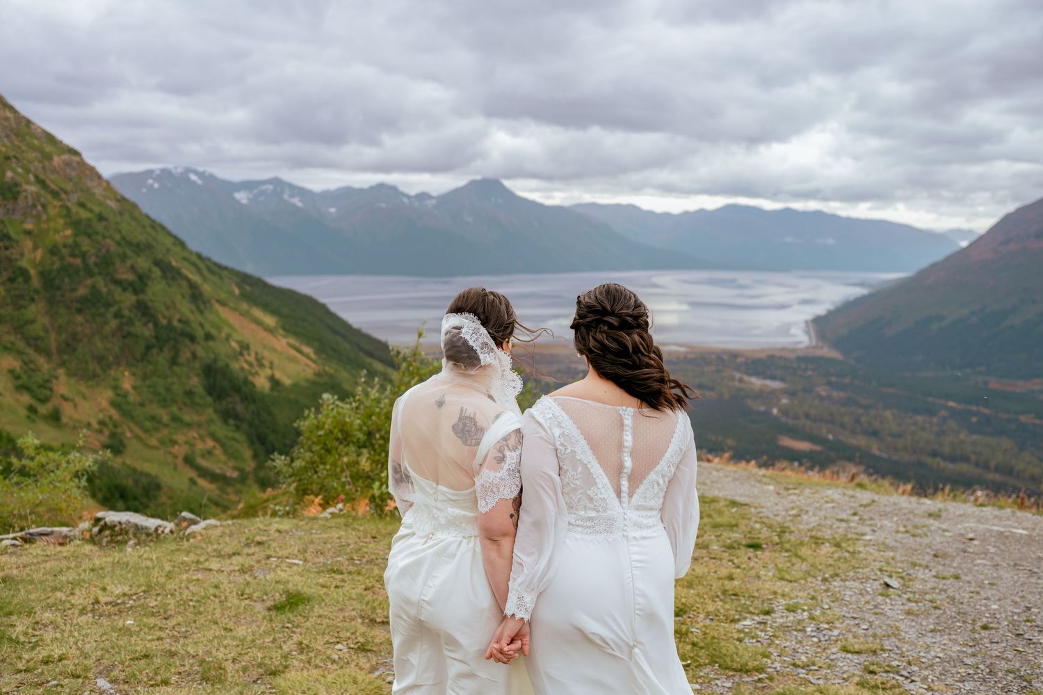 Two people in white wedding dresses hold hands while looking out over a mountain valley landscape in Alaska.
