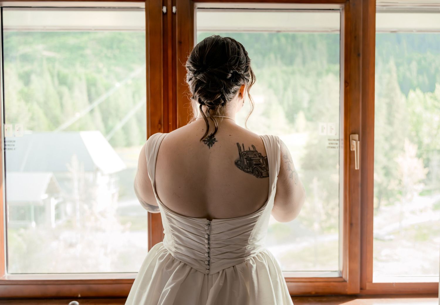 Bride in white wedding dress with back tattoo gazing out window at mountain views.