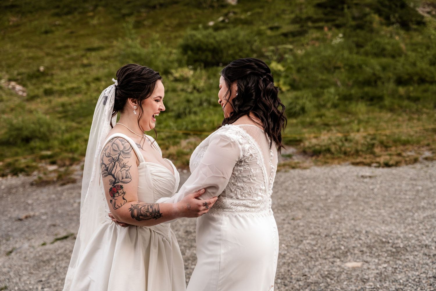 Two brides in white wedding dresses share a joyful moment outdoors, one with tattooed arms, smiling at each other.