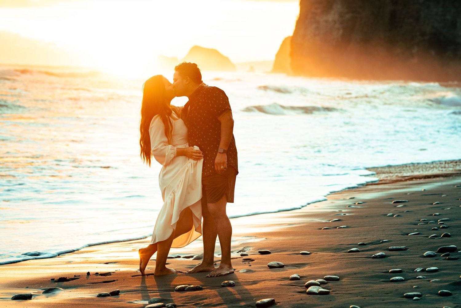 Romantic couple kissing on a sandy beach at golden sunset with ocean waves and dramatic cliffs in background.