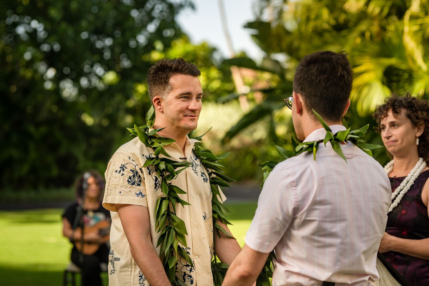 Two grooms exchange vows outdoors wearing floral leis at a tropical Hawaiian wedding ceremony.