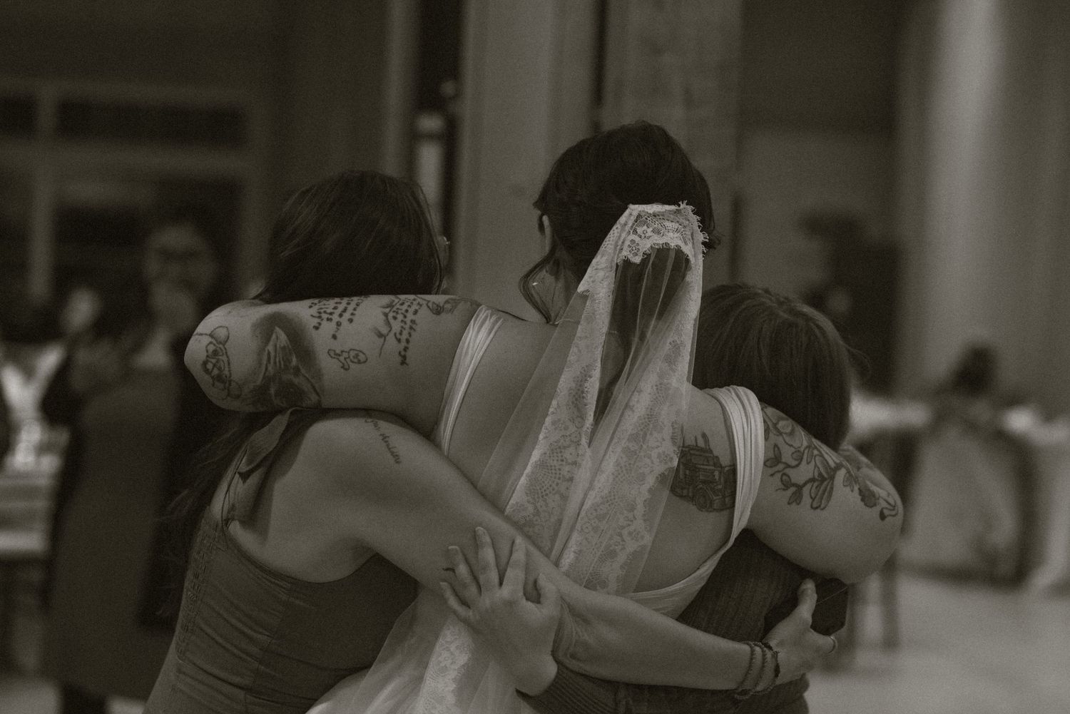 Black and white photo of a bride in veil hugging tattooed bridesmaids in a group embrace at wedding reception.