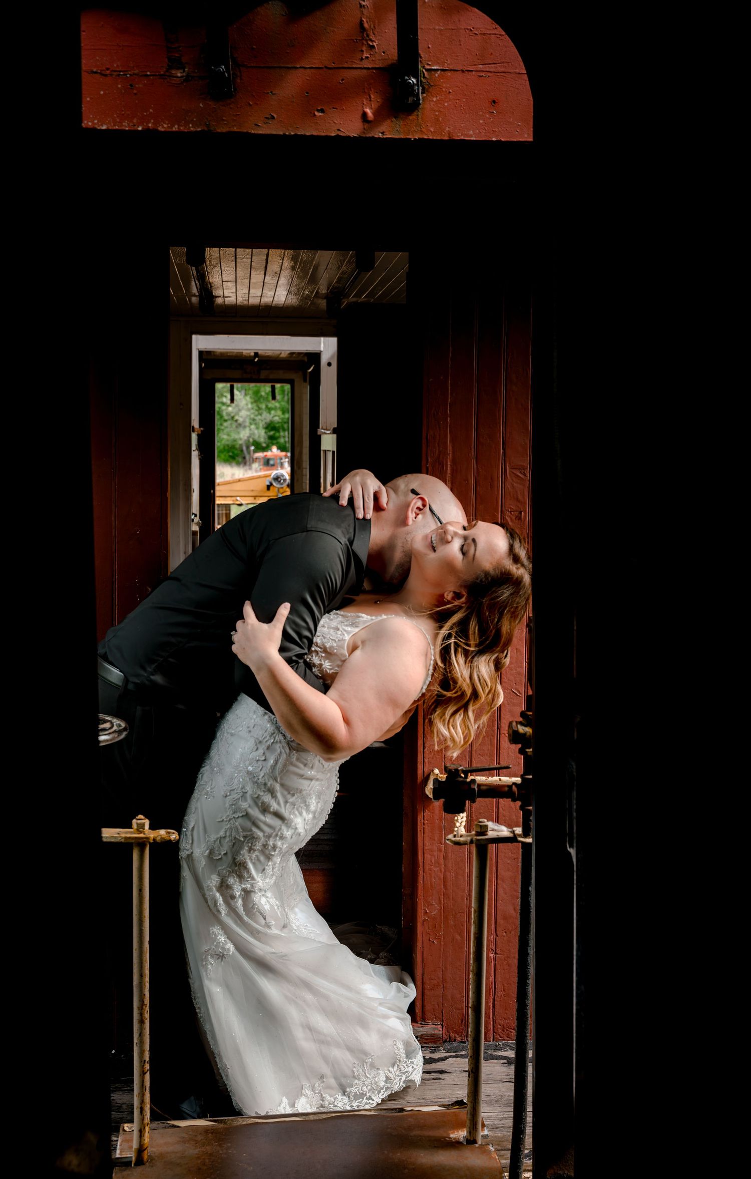 Bride and groom share a romantic dip kiss in a rustic red train car doorway on their wedding day.