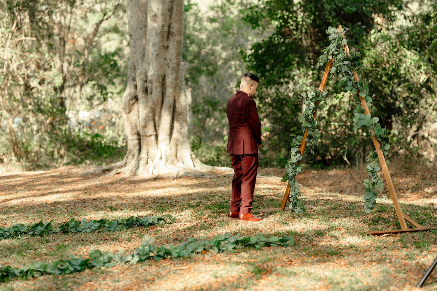 Groom in burgundy suit stands beneath floral arch in sunlit forest clearing during outdoor wedding ceremony.