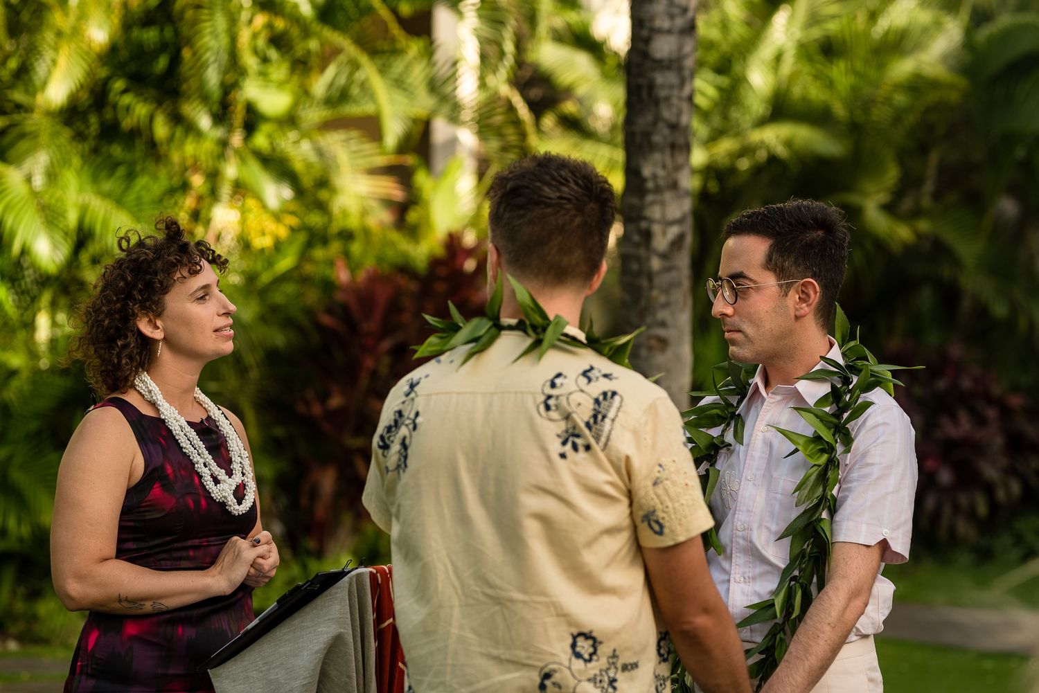 Two men in floral shirts and lei garlands exchange vows with an officiant outdoors in a tropical Hawaiian setting.