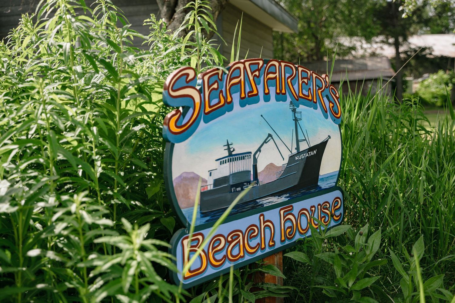 Seafarer's Beach House in Homer Alaska wooden sign surrounded by lush green foliage with a fishing boat illustration.