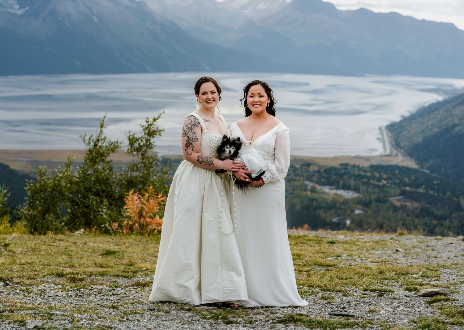 Two brides in white gowns smile together on a mountain overlook with scenic river valley and misty peaks behind them.