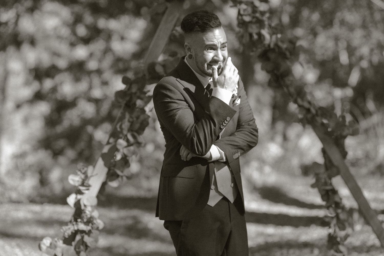 Man in suit laughing outdoors in black and white wedding photo among trees.