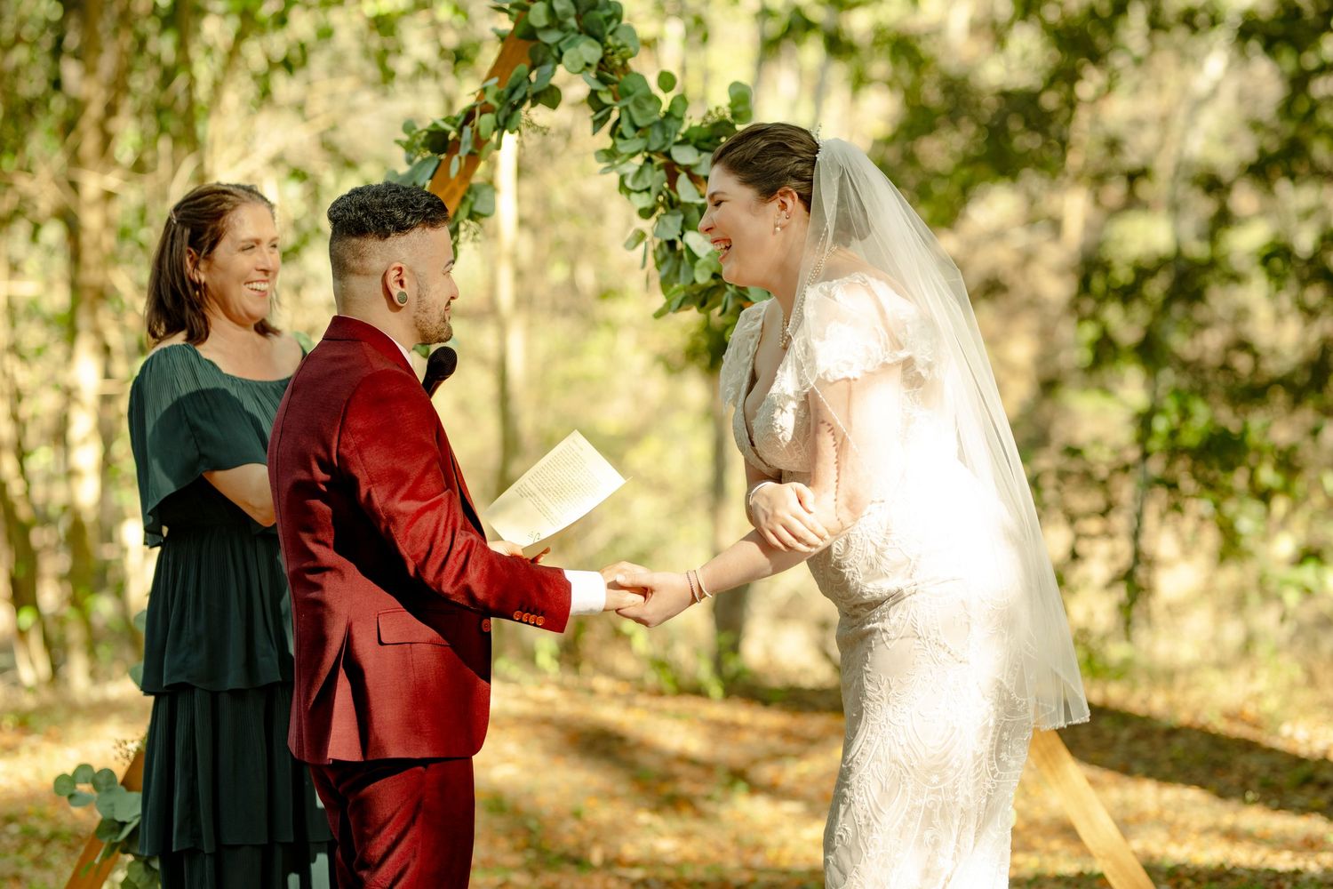 Outdoor wedding ceremony in forest with bride in white gown, groom in red suit, exchanging vows before officiant.