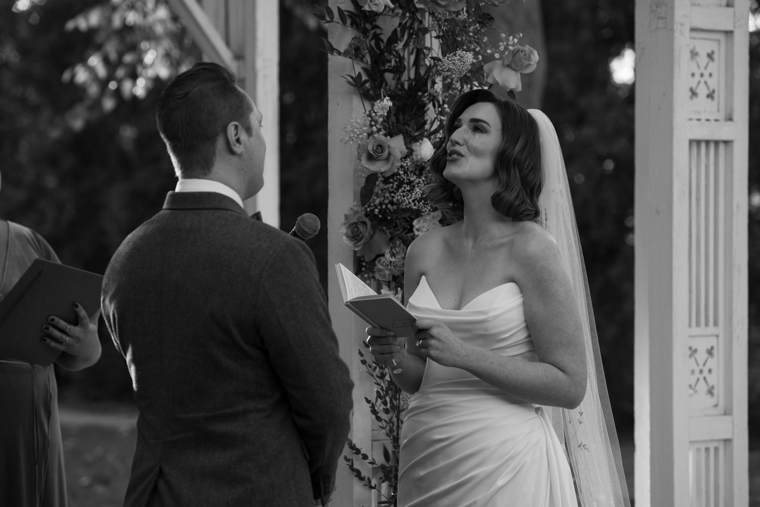 Black and white photo of a bride reading vows to her groom during an outdoor wedding ceremony beneath a floral arch with tears falling.